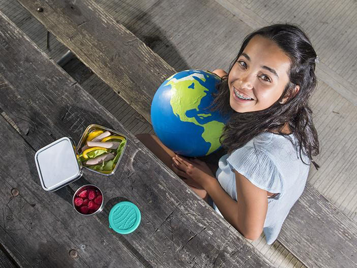 Break free from plastics image of a girl holding a globe next to a table of ECOlunchbox snack containers.