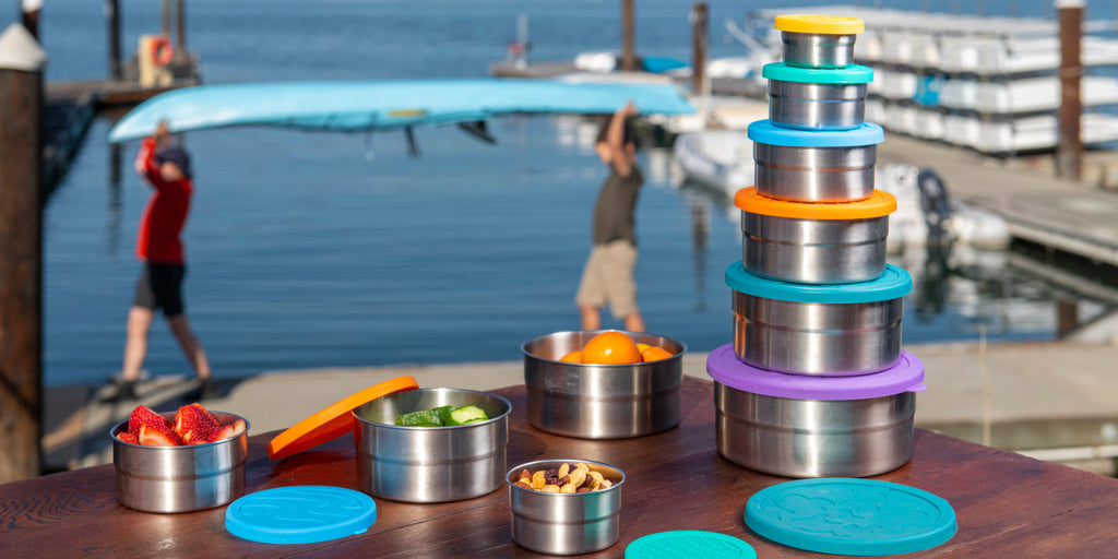 Several types of Blue Water Bento stainless steel lunchboxes on a wooden table near a pier with people carrying a canoe in the background.